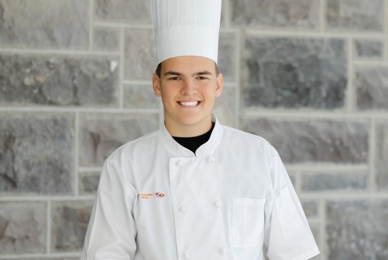 Headshot of Will Racek wearing a chef's hat and chef's uniform and standing in front of a Hokie Stone wall.