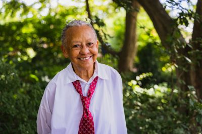 Nikki Giovanni smiling for a photo wearing a white shirt and red tie. Trees and other greenery are in the background. 