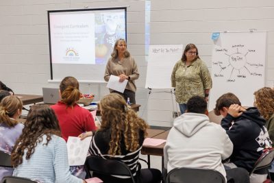 Two teachers from Rainbow Riders child care present to a group of high schoolers with a presentation, white board, and easel.