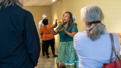Vicky Ferguson, in a green Monacan-style dress and donning a feather in her hair, guides visitors through an art exhibit for Indigenous Peoples' Day 2023.