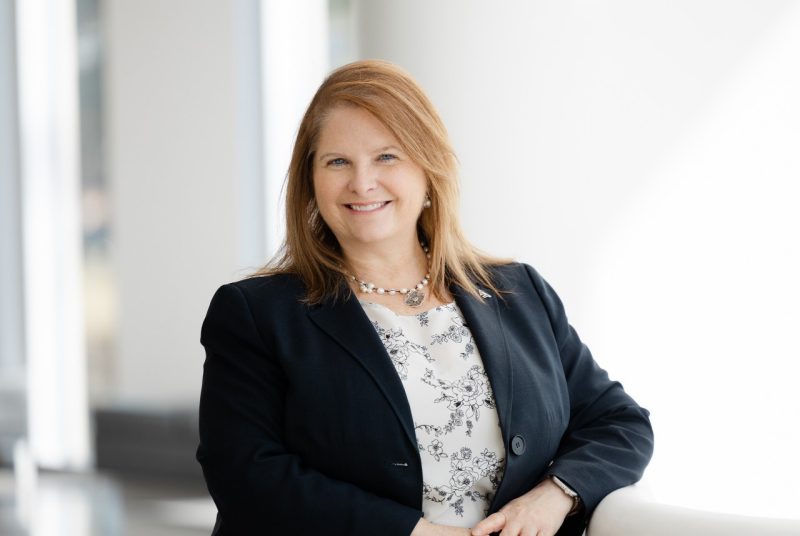 Headshot of Christine Watkinson wearing a business jacket and floral necklace and resting her elbow on a ledge.