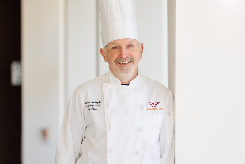 Mike Arrington smiles and poses for a photo wearing a white chef's apron and a toque blanche.