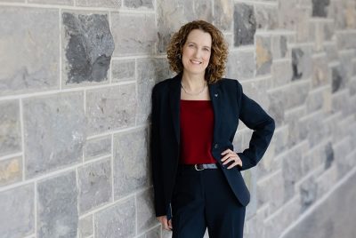 Amy Sebring, wearing a suit, leans against a stone wall and smiles with a hand on her hip.