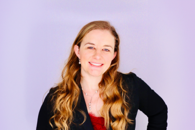 Rachel miles, dressed in a suit, stands in front of a light, blank background.