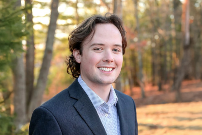 Portrait shot of Alexander Efird, wearing a suit without a tie, standing outside with a semi-autumnal background.