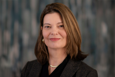 Portrait of Sharon Pit in a dark suit and pearl necklace in front of a unfocused background.