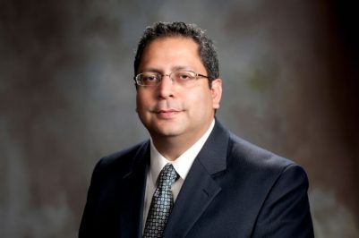 Guru Ghosh in a suit and tie in front of a portrait background.