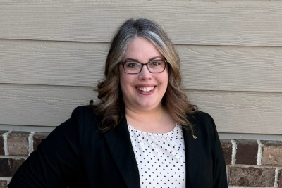 Rebecca Bott-Knutson stands and smiles against a wood and brick wall, wearing a blazer with a VT pin.
