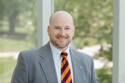 Portrait shot of Brennan Shepard in a suit and Hokie-colors tie, standing in front of a window